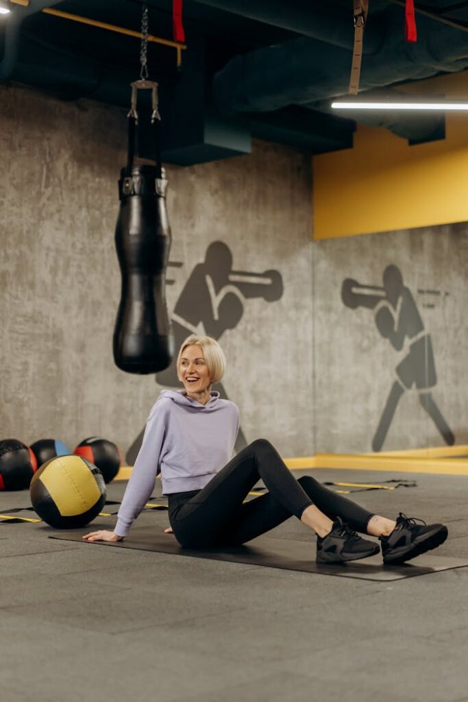 A woman relaxes on a gym floor surrounded by boxing and fitness equipment, smiling warmly.